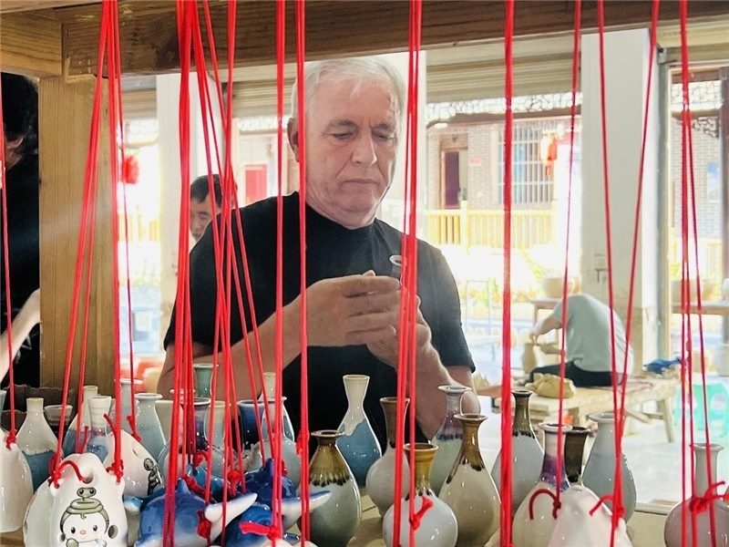鄉(xiāng)愁如同一根根紅線，將游子與故鄉(xiāng)永遠(yuǎn)地牽連到一起?；逄账囄幕瘎?chuàng)意一條街的母氏陶藝館內(nèi)，紅線吊著陶瓶，思緒游走萬里。摩爾多瓦“信息標(biāo)簽”通訊社社長亞歷山大·塔納斯端詳著館內(nèi)展示的陶藝作品，并挑選出心儀的小擺件帶回摩爾多瓦，也帶走花茂村的“鄉(xiāng)愁”。（張亞欣攝）